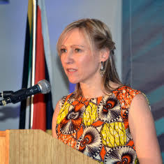 White woman at a podium speaking into the microphone. She wears a multicolored shirt with a vibrant orange and yellow design.