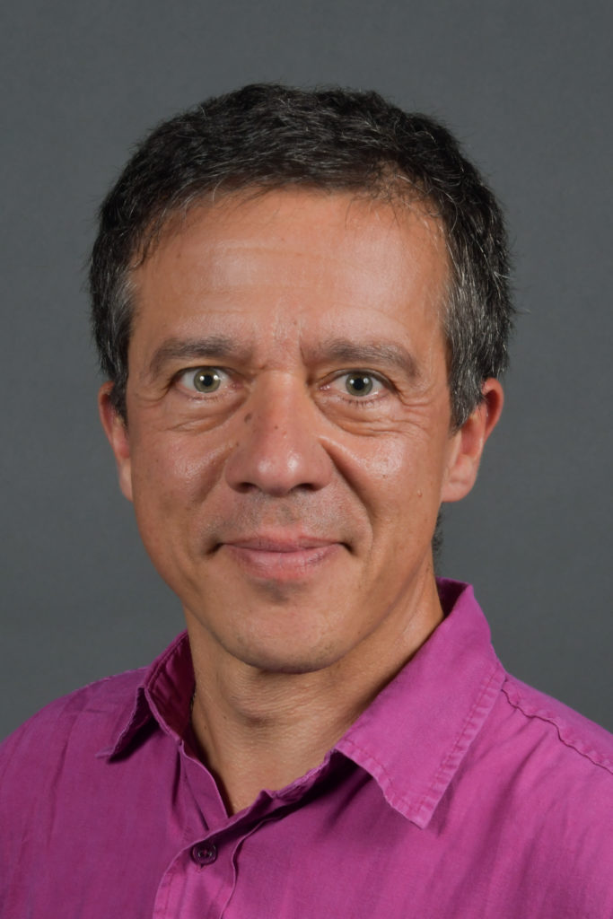 Light brown skinned man with dark brown/blackish hair and a fuscia shirt with collar smiles with lips closed.