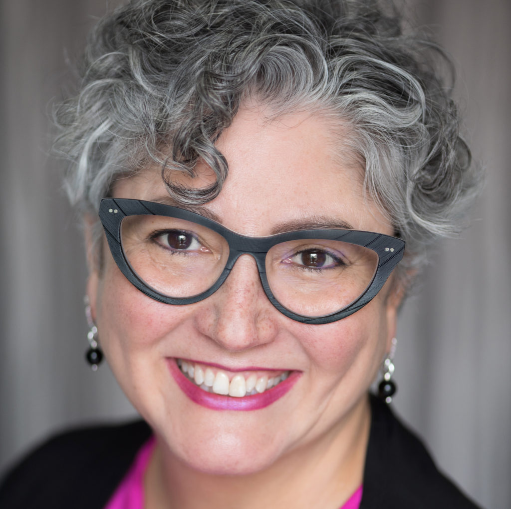 femme aux cheveux bouclés gris et lunettes souriant, devant un fond gris.