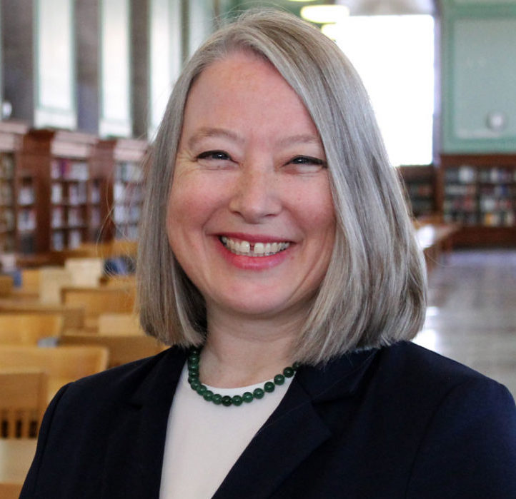 Uma mulher sorridente, com cabelo cinza claro na altura dos ombros, camisa branca e blazer preto e colar azul. Ela está em uma biblioteca.